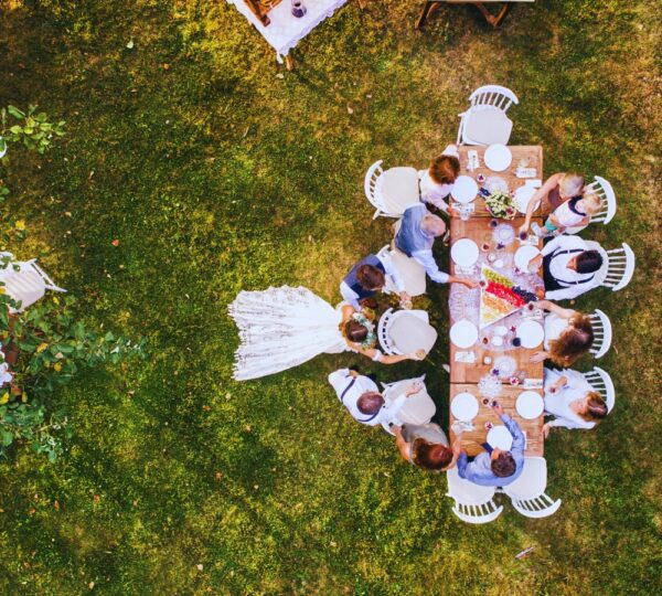 Stock image of a wedding couple eating at a long table outside with guests 10 guests. Bride and groom are standing facing table.