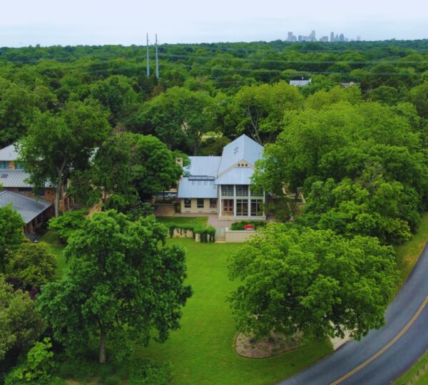 Aerial Shot of back of a large single family tan home, solar panels, light roof surrounded by large green trees agains the backdrop of Dallas Skyline.